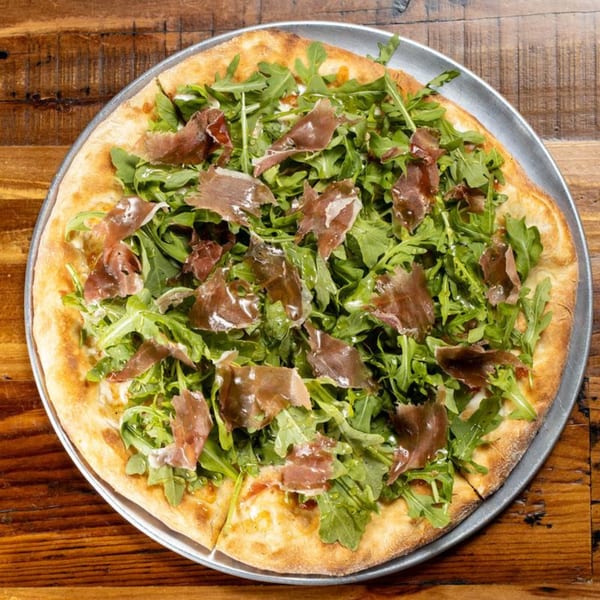 The Brick Oven Pizza's prosciutto and arugula pizza on a metal tray, on a wooden table.
