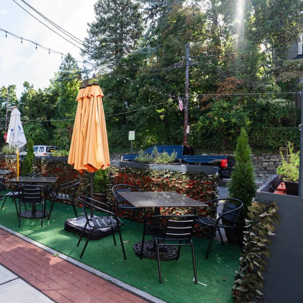 Outdoor patio of The Brick Oven Pizza with black tables, chairs, and orange umbrellas on green turf.